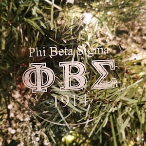 May include: Clear, round Christmas ornament with the Greek letters Phi, Beta, and Sigma, and the year 1914. The words "Phi Beta Sigma" are above the letters. The ornament hangs from a sheer ribbon against a backdrop of a Christmas tree.