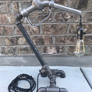 May include: A black and silver industrial style table lamp made from repurposed metal pipe fittings and a vintage gas pump nozzle. The lamp has a black cord and a clear light bulb.