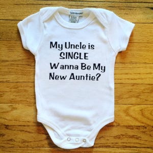 Puede incluir: Body de bebé blanco con el texto negro "My Uncle is SINGLE Wanna Be My New Auntie?". El body tiene mangas cortas y corchetes en la parte inferior. La marca "TEDDYBOOM" está impresa en la etiqueta.