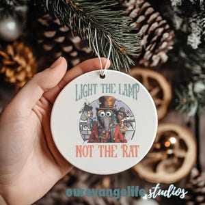 May include: A white, circular Christmas ornament featuring the text "LIGHT THE LAMP NOT THE RAT" and a cartoon illustration of a character in a top hat and red coat. The ornament is held by a hand.