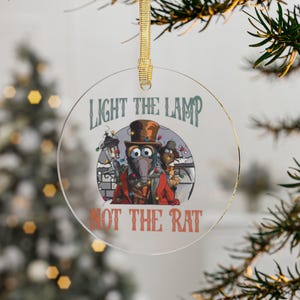 May include: A clear, round Christmas ornament with a gold ribbon. The ornament features a cartoon image of two characters in festive attire, with the text "LIGHT THE LAMP NOT THE RAT" in green and orange.