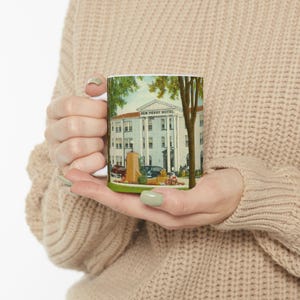 May include: White ceramic mug featuring a vintage illustration of the New Perry Hotel. The image shows a light blue building with white columns, trees, and a vintage car. The mug is held by a person wearing a beige sweater.