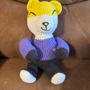 Peut inclure: Une poupée ours en crochet faite à la main avec une tête jaune, un visage blanc, un pull violet, un pantalon noir et des pieds blancs. L'ours a un visage souriant avec des yeux noirs et un nez noir. L'ours est assis sur une surface marron.