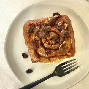 Puede incluir: Un solo rollo de canela con nueces pecanas y pasas en un plato blanco con un tenedor negro. El rollo está glaseado y tiene un color marrón dorado.
