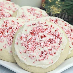 May include: A plate of frosted sugar cookies topped with crushed peppermint candy. The cookies have a white frosting base and are arranged on a white rectangular plate. The background includes greenery and a red and black plaid cloth.
