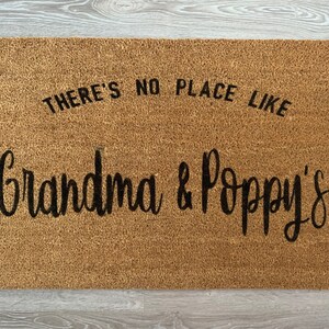 May include: A brown coir doormat with the text "There's no place like Grandma & Poppy's" in black lettering.