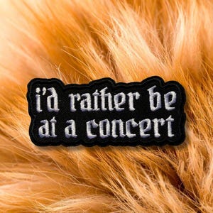 May include: A black embroidered patch with a jagged, irregular border. The text "i'd rather be at a concert" is stitched in a white, gothic-style font across the centre of the patch, creating a high-contrast design with clear, legible lettering.
