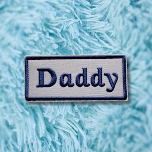May include: A rectangular embroidered patch featuring a light grey background with a dark blue border. The word Daddy is stitched in dark blue thread in a serif font across the centre of the patch, which has a clean, finished edge.