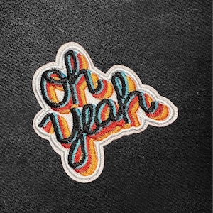 Puede incluir: Parche bordado con la frase "oh yeah" en una fuente retro. El texto es negro, con acentos superpuestos en naranja, rojo y azul. El parche tiene un borde blanco y está sobre un fondo negro.
