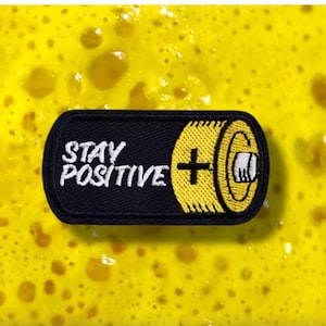 Könnte beinhalten: Schwarzer rechteckiger Aufnäher mit den weißen Worten "STAY POSITIVE". Eine gelbe Batterie-Grafik mit Pluszeichen befindet sich auf der rechten Seite. Der Aufnäher liegt auf einem strukturierten gelben Hintergrund.