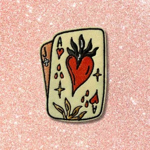 May include: A playing card patch featuring the Ace of Hearts and Jack of Hearts. The Ace of Hearts has a large red heart with a black flame design above it. The card has gold accents, including stars and leaves, with a black border.