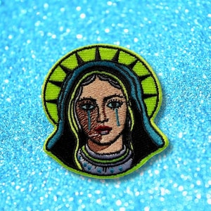 May include: An embroidered patch featuring a stylised portrait of a figure with a tan face, red lips, and blue tears. The figure wears a dark blue head covering with a bright green halo border, all outlined in black thread on a circular shape.