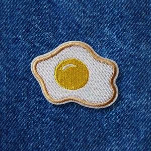 Puede incluir: Parche bordado con un diseño de huevo frito. El huevo tiene un fondo blanco con una yema amarilla y un borde dorado. El parche está sobre un fondo de mezclilla azul.