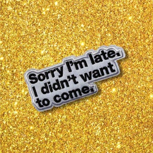 May include: A rectangular embroidered patch with a light grey background and a dark grey border. The patch features the black embroidered text "Sorry I'm late. I didn't want to come." in a bold, sans-serif font, arranged in three distinct lines.