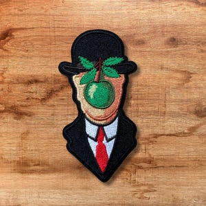 Peut inclure: Patch brodé représentant un homme portant un chapeau melon noir avec une pomme verte qui lui couvre le visage. L'homme porte un costume noir et une cravate rouge.