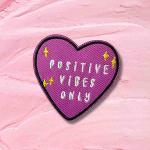 May include: A heart-shaped patch in shades of purple with the words "POSITIVE VIBES ONLY" embroidered in white. The patch is decorated with yellow starbursts. The patch is on a pink background.