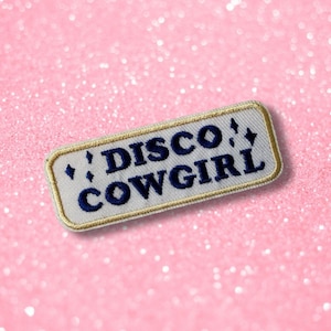 Może przedstawiać: Prostokątna haftowana naszywka z jasnoszarym tłem i złotą obwódką. Napis DISCO COWGIRL jest wyhaftowany ciemnoniebieskimi wielkimi literami, w towarzystwie małych ciemnoniebieskich czteroramiennych gwiazdek rozmieszczonych wokół tekstu.