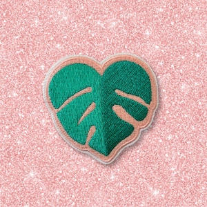 May include: A green embroidered monstera leaf in the shape of a heart, on a pink glitter background.