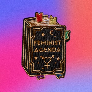 Puede incluir: Un parche bordado negro con ribetes dorados que representa un libro cerrado con el texto "C. Feminist Agenda" y un símbolo de género. El libro tiene un marcapáginas dorado y algunos otros elementos decorativos.