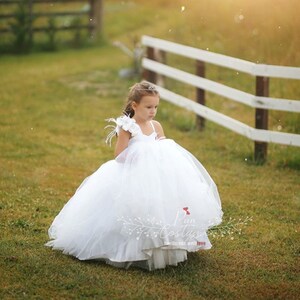 May include: A young girl wearing a white tulle dress with a single white flower and feather on the shoulder. The dress has a sweetheart neckline and a full skirt. The girl is standing in a grassy field with a white fence in the background.