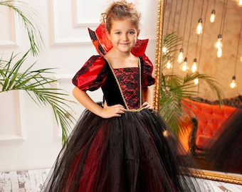 May include: A young girl wearing a red and black princess costume with a tulle skirt and a lace bodice. The costume has a gold trim and a red cape. The girl is smiling and looking at the camera.