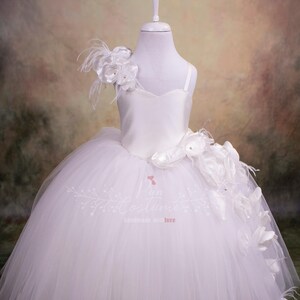 May include: White tulle dress with a satin bodice and a large white flower on the shoulder. The dress has a full skirt and is perfect for a special occasion.