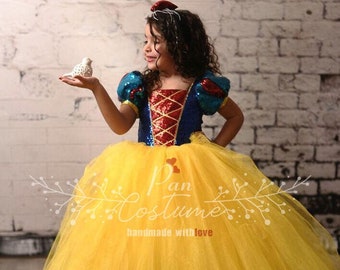 Puede incluir: Una niña pequeña con un disfraz de Blancanieves. El vestido es azul con detalles rojos y una falda de tul amarilla. Está sosteniendo una figurita de pájaro blanca.