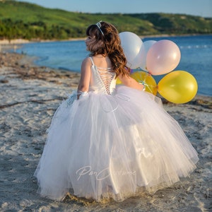 May include: A young girl in a white tulle dress and a silver tiara holds a bunch of colorful balloons on a sandy beach.