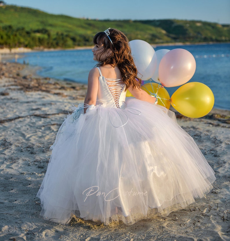 May include: A young girl in a white tulle dress and a silver tiara holds a bunch of colorful balloons on a sandy beach.