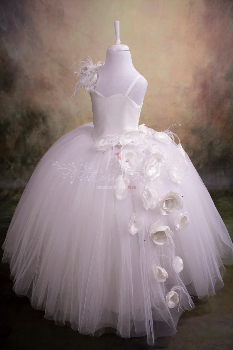May include: White tulle dress with a satin bodice and a single strap. The dress has a large white flower embellishment on the side.
