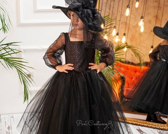 May include: A young girl dressed as a witch for Halloween. She is wearing a black tulle dress with a black velvet bodice and a black witch hat with a veil. The dress has a sparkly design. The hat has a black feather and a black bow.