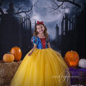 Girl posing in her Snow White dress with a big smile on her face, standing, holding her skirt with one hand. Orange and white pumpkins are placed on hay piles. A full moon partly covered with dry tree branches and iron gates are at the background.