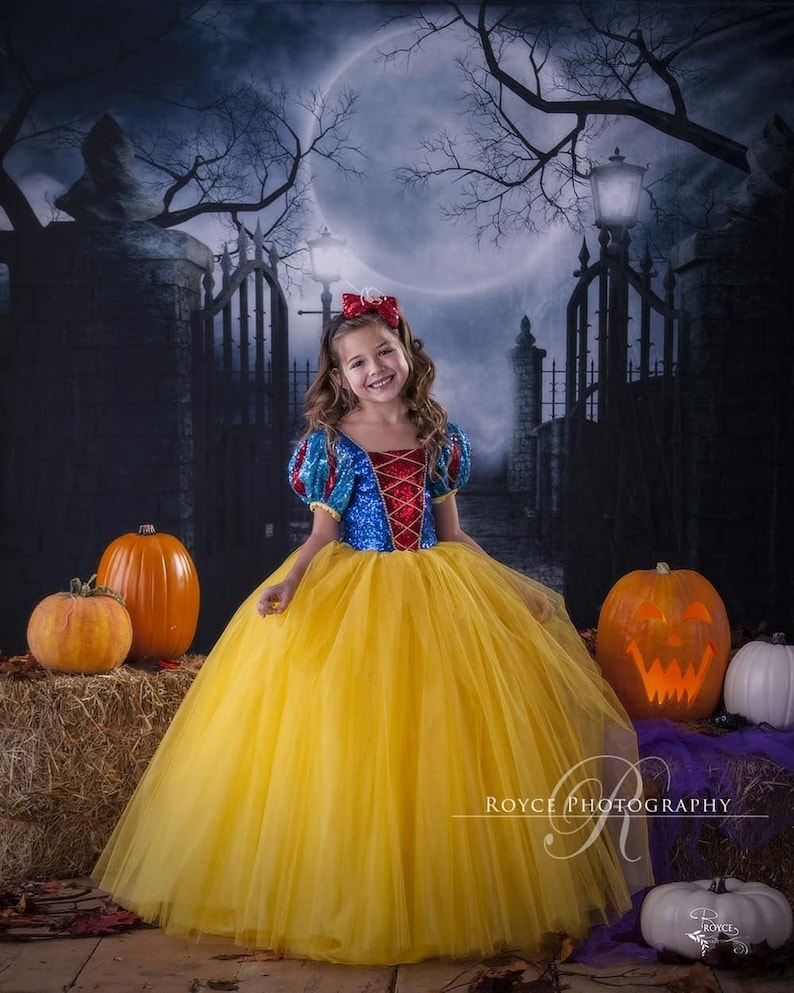 Girl posing in her Snow White dress with a big smile on her face, standing, holding her skirt with one hand. Orange and white pumpkins are placed on hay piles. A full moon partly covered with dry tree branches and iron gates are at the background.