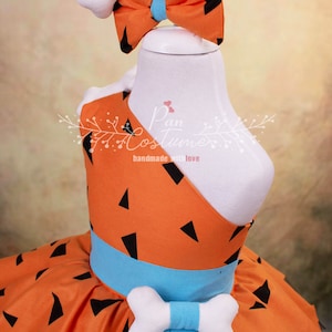 May include: Orange and black patterned dress with a blue sash and a white bone-shaped bow. The dress has a white bone-shaped headband with a blue bow.