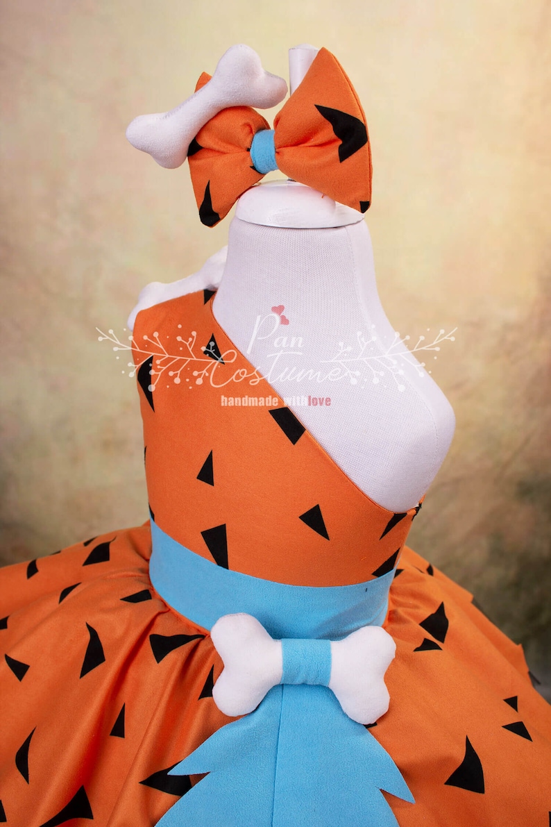 May include: Orange and black patterned dress with a blue sash and a white bone-shaped bow. The dress has a white bone-shaped headband with a blue bow.