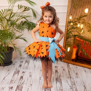 May include: A young girl wearing a bright orange and black costume with a blue bow and a bone-shaped belt. The costume is inspired by the cartoon character Pebbles Flintstone.
