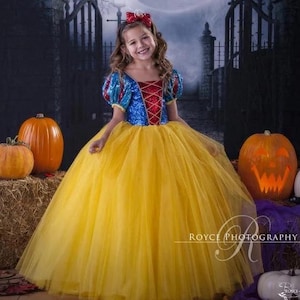 May include: A young girl dressed as Snow White in a blue and red sequined bodice and yellow tulle skirt. She is wearing a red bow in her hair and is standing in front of a backdrop of a gate and pumpkins.