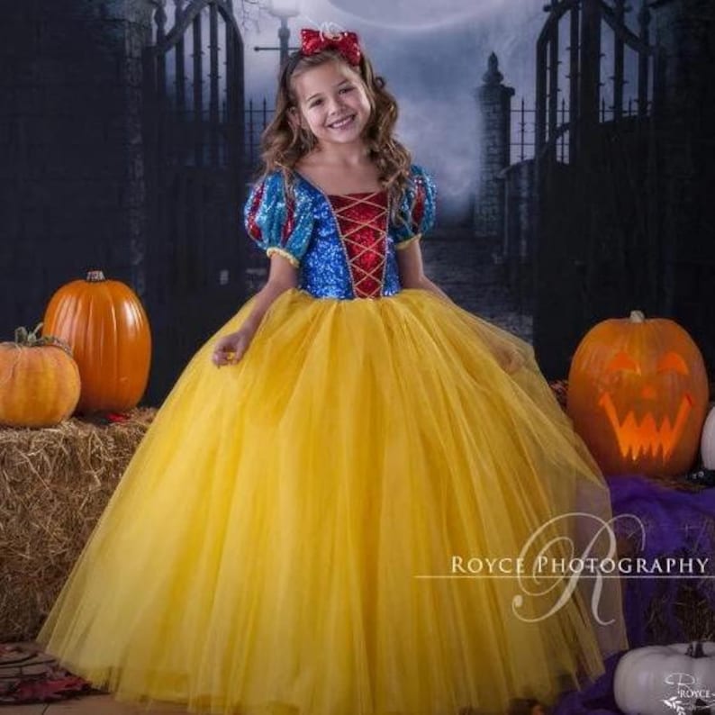 May include: A young girl dressed as Snow White in a blue and red sequined bodice and yellow tulle skirt. She is wearing a red bow in her hair and is standing in front of a backdrop of a gate and pumpkins.