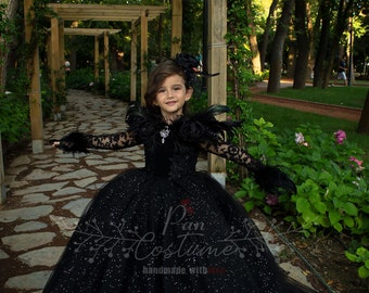 May include: A young girl wearing a black tulle dress with silver glitter and black feather accents. The dress has long sleeves and a high neckline. The girl is standing in a garden setting with a wooden pergola behind her. The text "Pan Costumes handmade with love" is visible on the dress.
