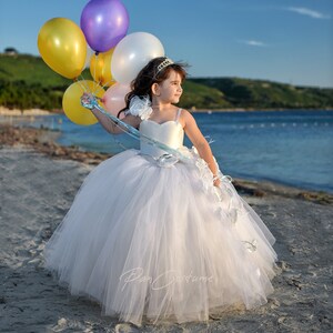 May include: A young girl in a white tulle dress with a blue sash and flower accents holds a bunch of colorful balloons on a beach.