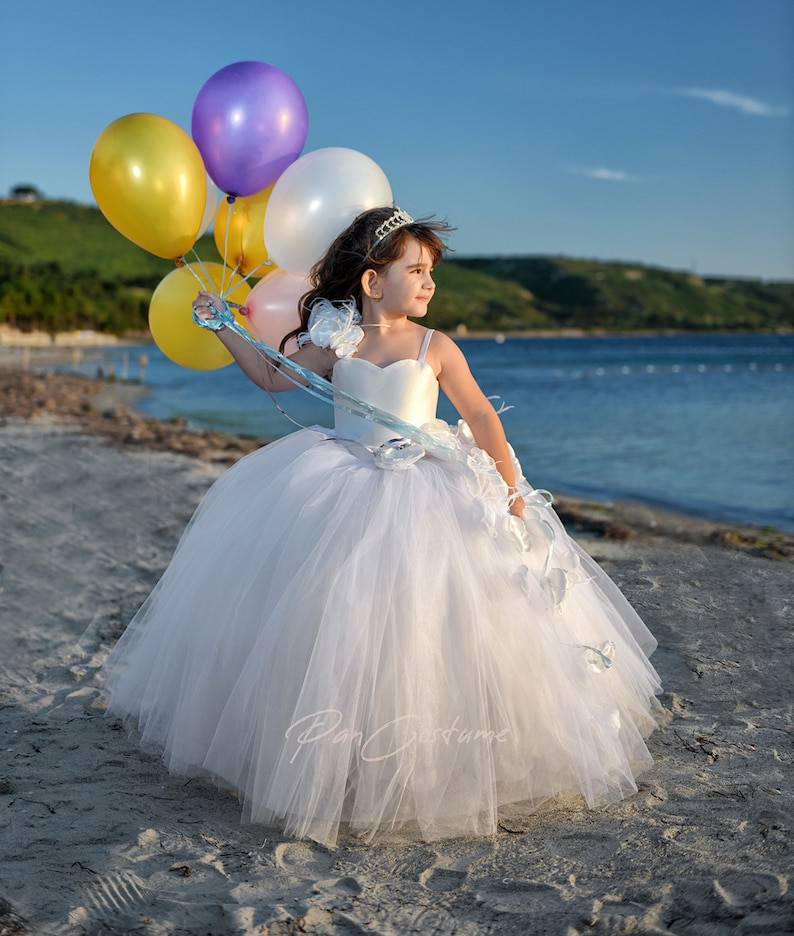 May include: A young girl in a white tulle dress with a blue sash and flower accents holds a bunch of colorful balloons on a beach.