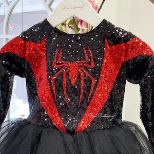 May include: Black and red sequined dress with a spider design on the front. The dress has long sleeves and a tulle skirt. The dress is on a hanger with the text "Pan Costume" on it.