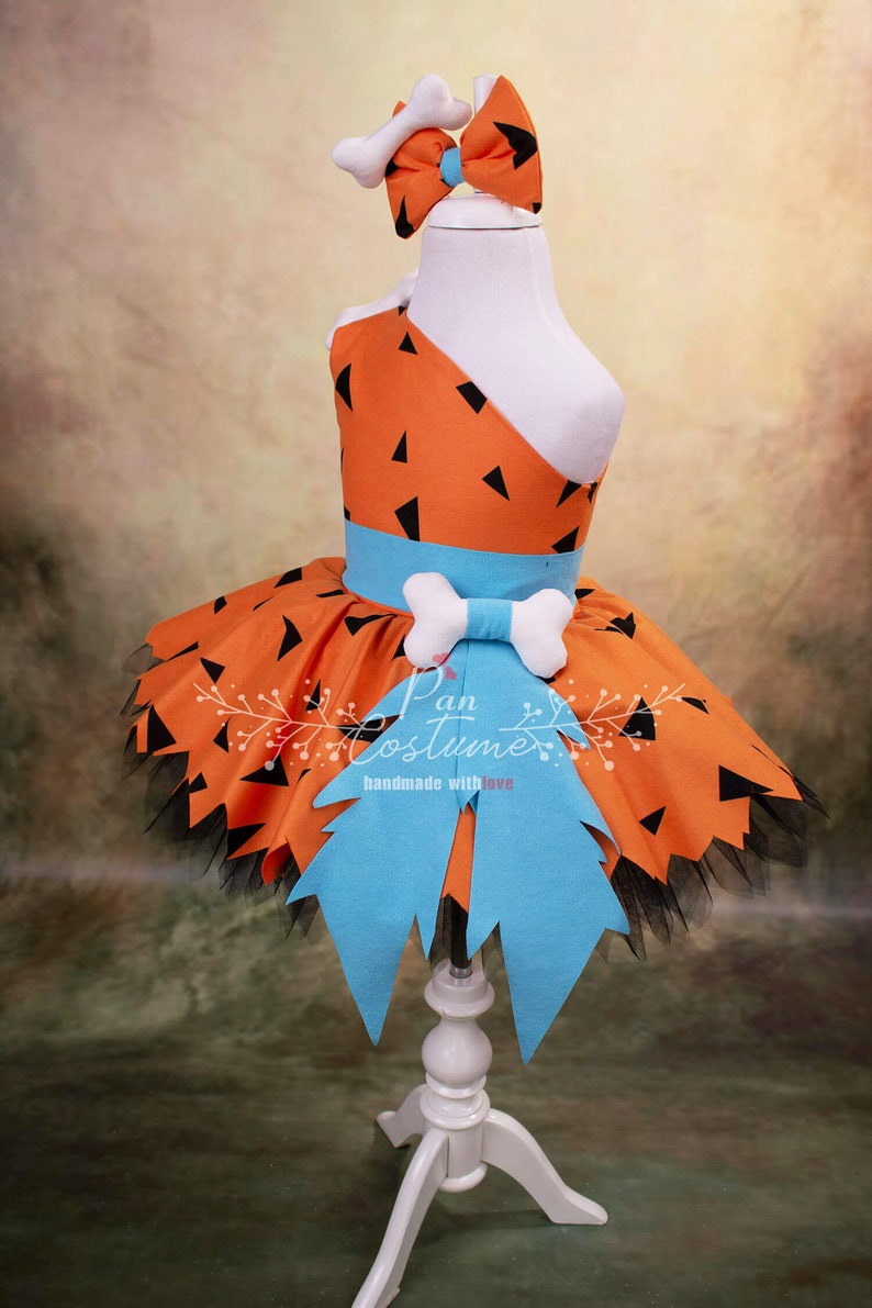May include: A child's costume featuring a bright orange and black dress with a blue sash and a bone-shaped bow. The dress has a tulle skirt and a jagged edge. The costume is on a white dress form.