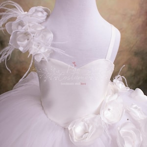 May include: White tulle dress with a satin bodice and white flower embellishments. The dress has a single strap and a large flower with feathers on one shoulder.