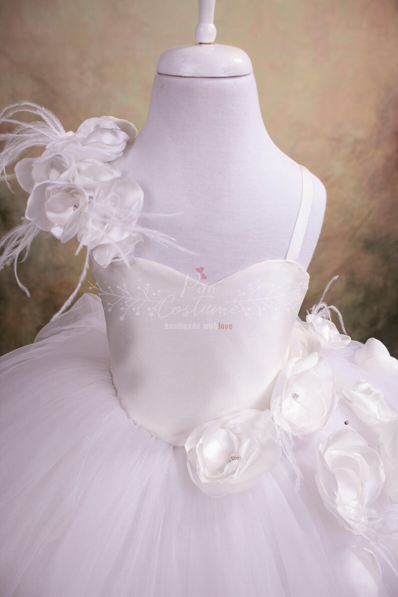 May include: White tulle dress with a satin bodice and white flower embellishments. The dress has a single strap and a large flower with feathers on one shoulder.