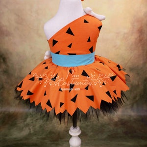 May include: Orange and black dress with a blue belt and a black tulle skirt. The dress has a one-shoulder design and is decorated with black triangles. The dress is on a white mannequin. The text "Pan Costume handmade with love" is on the dress.