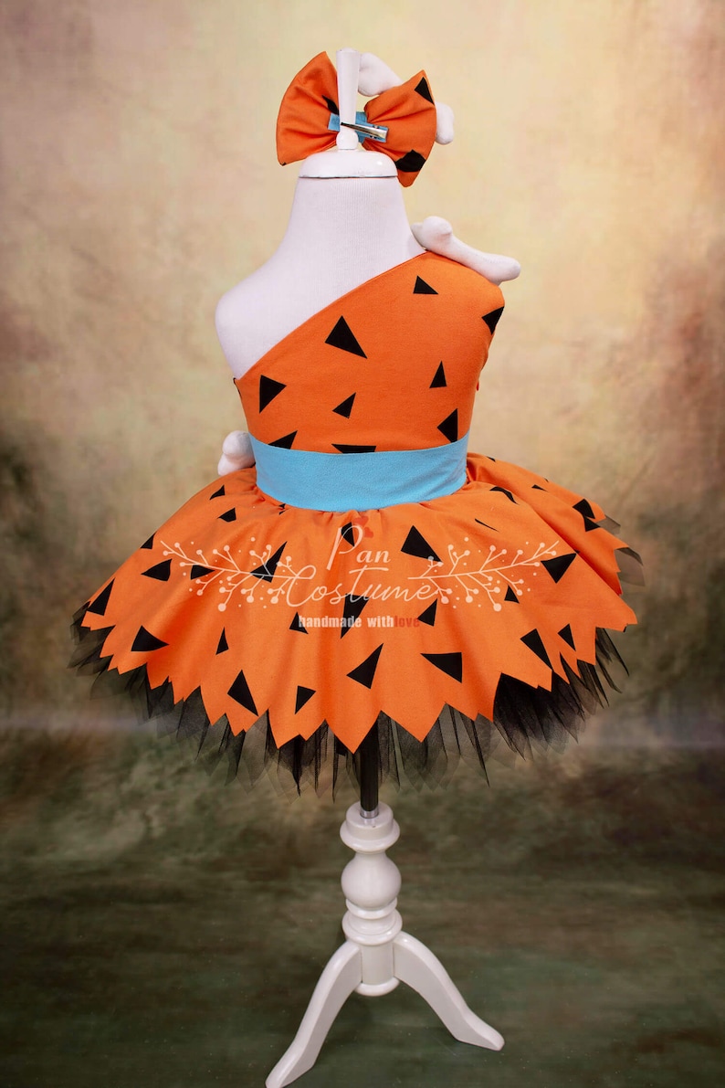 May include: Orange and black dress with a blue belt and a black tulle skirt. The dress has a one-shoulder design and is decorated with black triangles. The dress is on a white mannequin. The text "Pan Costume handmade with love" is on the dress.