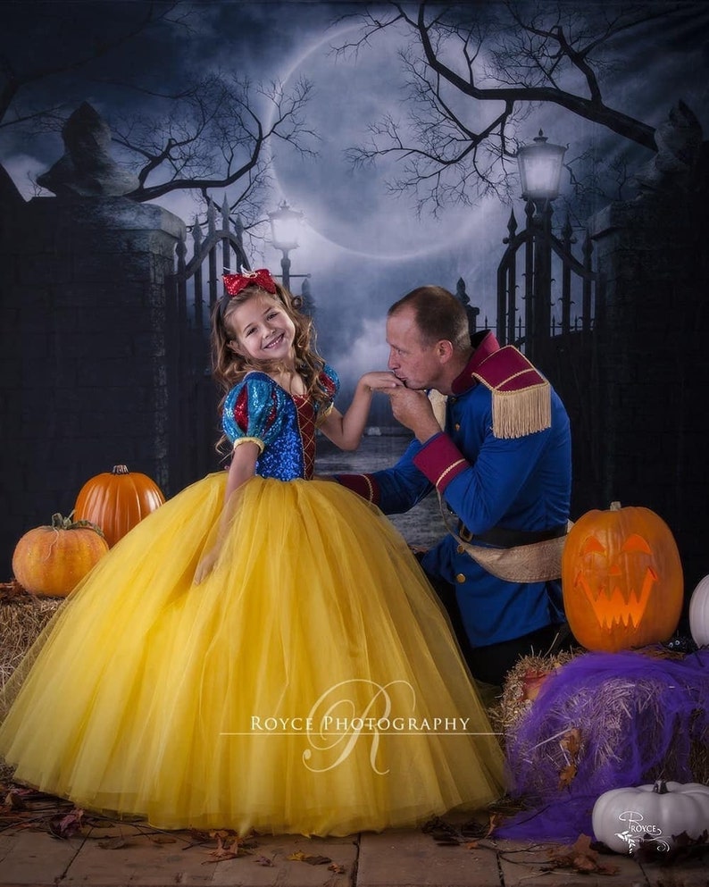 Sweet little girl in her fluffy Snow White costume, standing, her head tilted slightly with a cheerful smile on her face. Halloween decorations with pumpkins  and creepy background, her daddy disguised as her prince, kneeling down, kissing her hand.