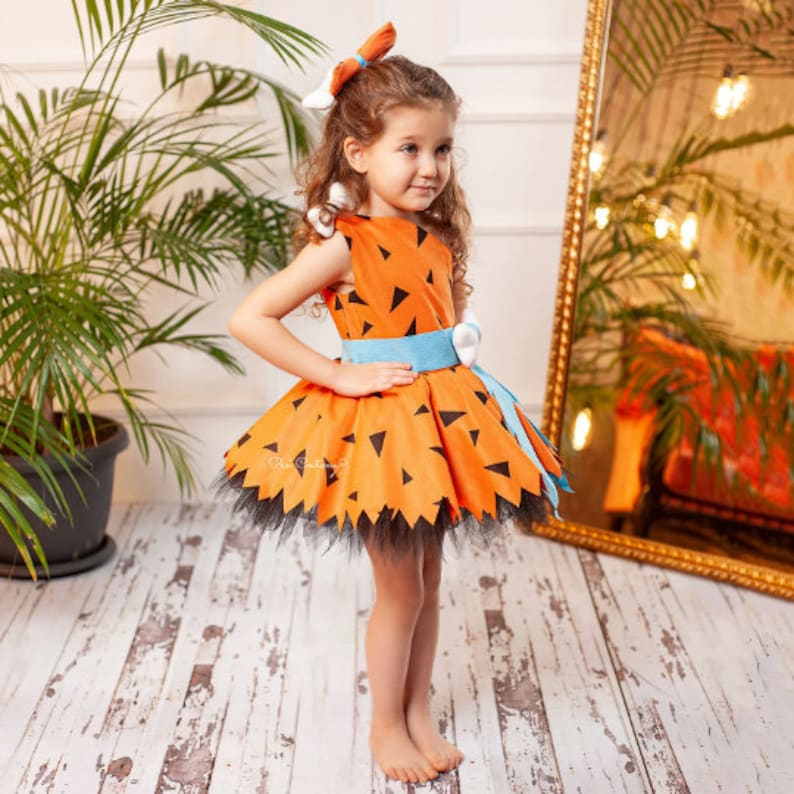 May include: A young girl wearing an orange and black dress with a blue belt. The dress has a pattern of triangles and is styled after the character Pebbles from the cartoon The Flintstones. The dress has a tulle skirt and a black tulle trim.