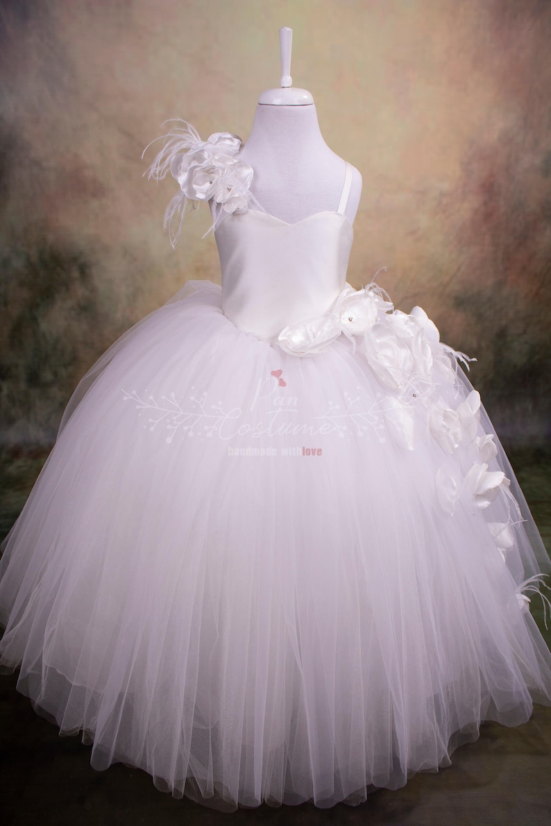 May include: White tulle dress with a satin bodice and a large white flower on the shoulder. The dress has a full skirt and is perfect for a special occasion.